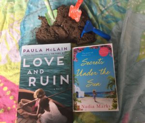 Love and Ruin and Secrets Under the Sun book sitting on a beach blanket in front of a pile of sand with shovels and rakes in bright colours.
