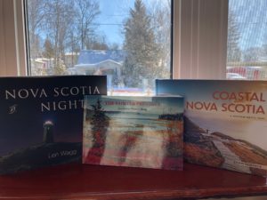Three books - Nova Scotia at Night, The Painted Province and Coastal Nova Scotia, in front of a window with the sun shining in and snow in the background